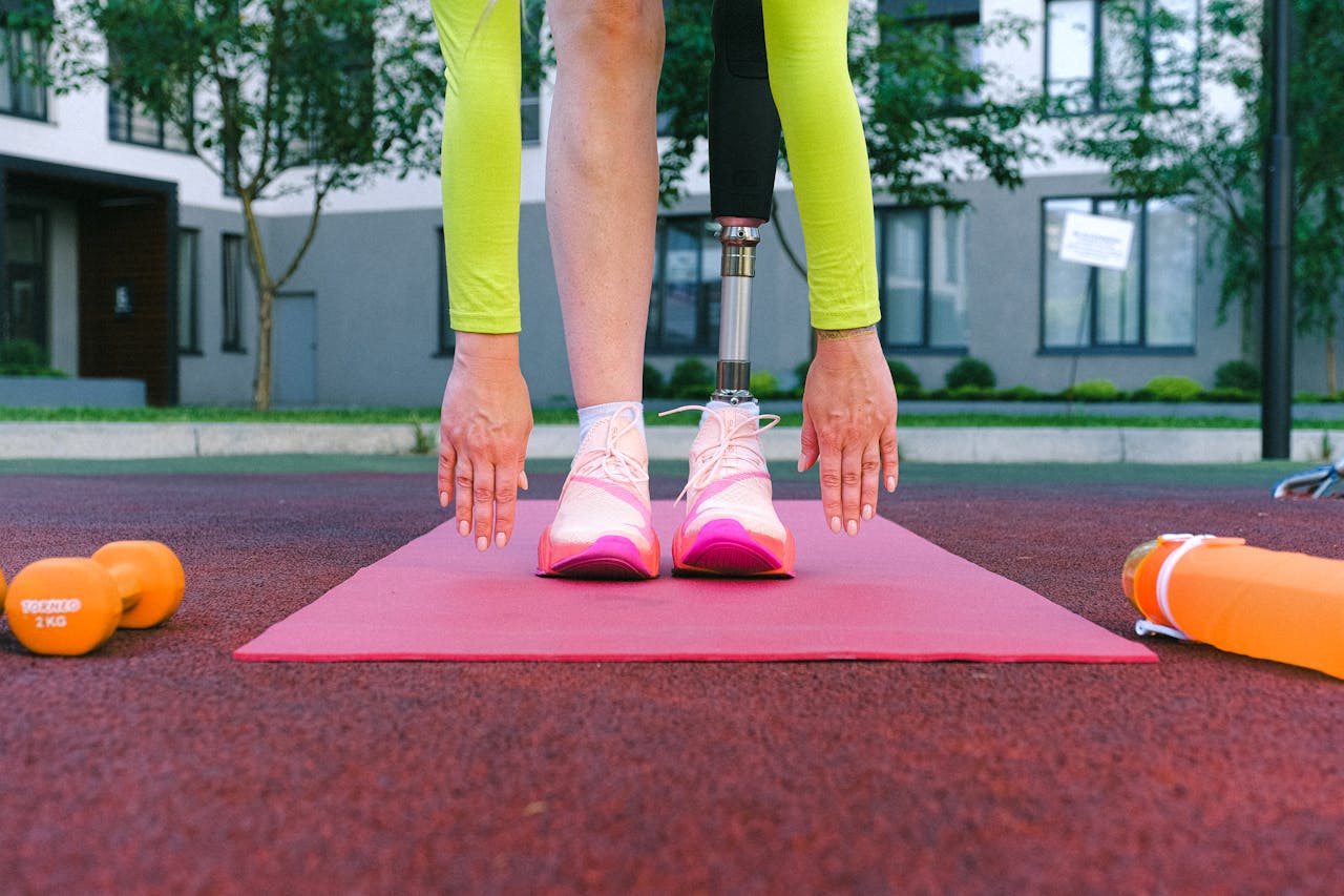 An adult stretching with a prosthetic leg on a red exercise mat outdoors.