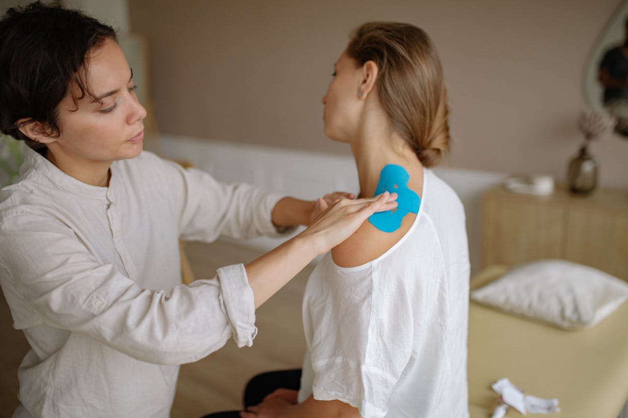 A physiotherapist applies blue kinesiology tape to a patient's shoulder for muscle support.